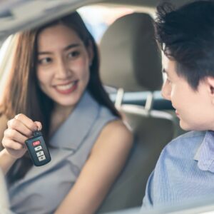 Asian woman showing car remote key to man friend sitting in vehicle at car rental company together