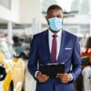Young Black Car Salesman Wearing Medical Mask Posing At Workplace In Showroom